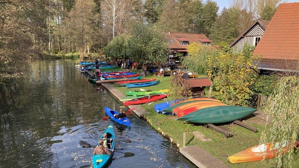 Une rivière paisible où sont amarrés des canoës le long de la rive, entourée d’arbres et bordée d’un petit bâtiment.
