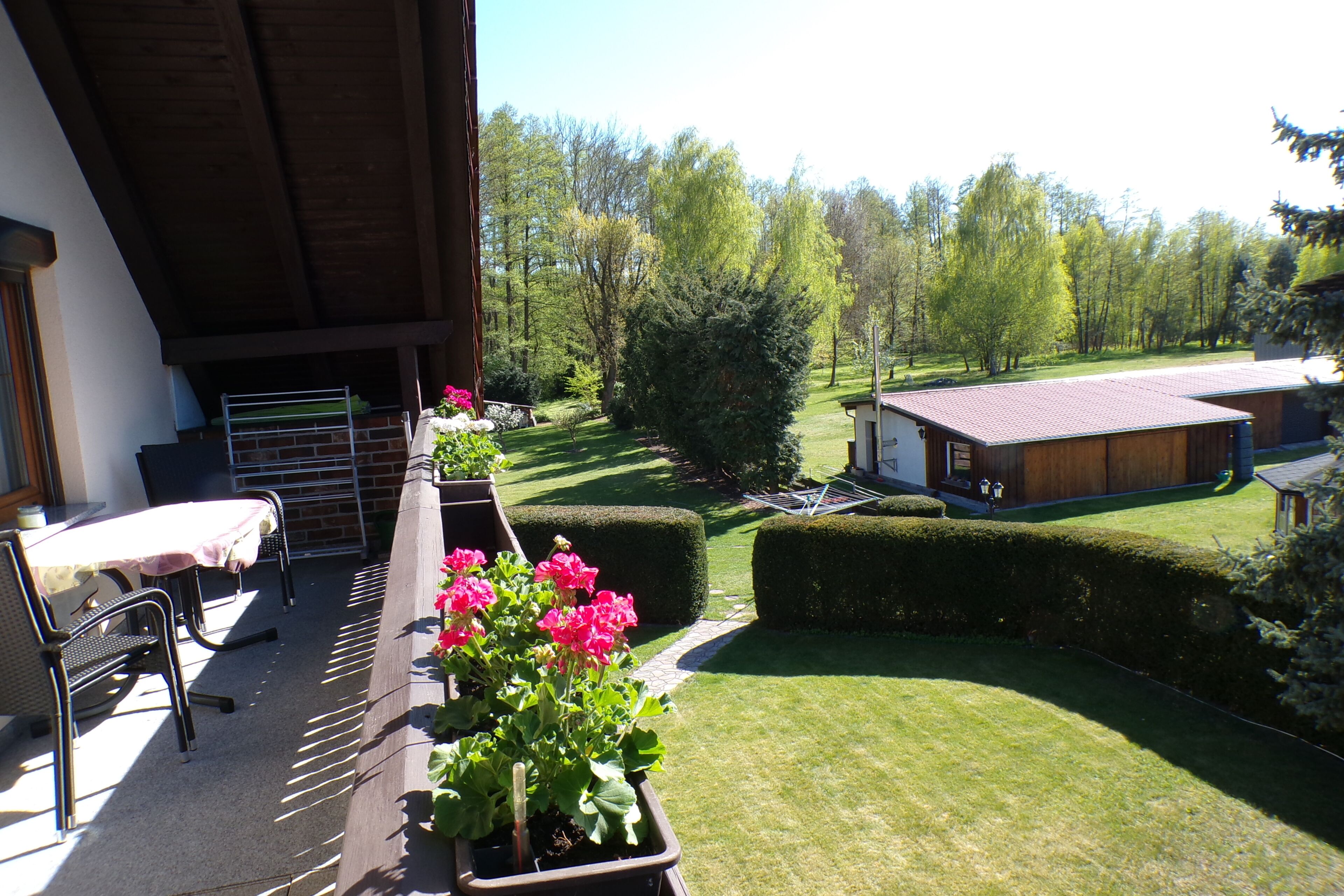 A balcony with a table and chairs, a potted plant with flowers, and a view of a well-maintained lawn and trees.