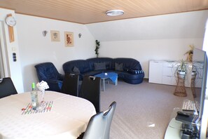 A room with a dining table, chairs, a sofa, a small table with a blue tablecloth, a white cabinet, and a plant.