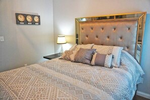 Un dormitorio con una cama grande, un cabecero capitonado, ropa de cama estampada, una mesita de noche con una lámpara y un reloj de pared que muestra varias zonas horarias.