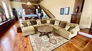A spacious living room with a large sectional sofa, a round coffee table, and a patterned rug. A staircase leads to an upper level, and the room features wooden flooring and a chandelier.