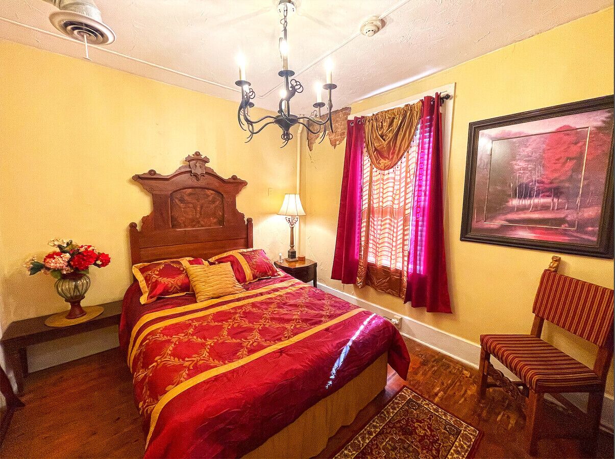 Un dormitorio con una cama grande, cabecera de madera, candelabro, mesita de noche con lámpara, una silla, ventana con cortinas e imagen enmarcada en la pared.