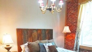 A bedroom with a wooden headboard, a bed with patterned bedding, a nightstand with a lamp, and a chandelier. - Shopping District Apt | Heart of Historic Downtown (Marietta)