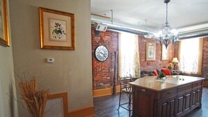 A spacious kitchen with a large island, a dining area with a brick wall, and a living room with a sofa and a chandelier. - Shopping District Apt | Heart of Historic Downtown (Marietta)
