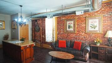 A living room with a brick wall, a wooden coffee table, a leather sofa with red cushions, and a kitchen area with a wooden island and a clock on the wall.