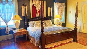 Una cama con dosel de madera y colcha estampada, flanqueada por dos mesitas de noche con lámparas, en una habitación con suelo de madera y cortinas en las ventanas.