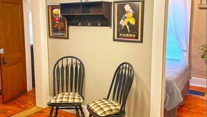Une pièce comprenant deux chaises en bois avec des coussins à carreaux, une étagère fixée au mur avec deux tableaux encadrés, un luminaire suspendu et une porte donnant sur une autre pièce.
