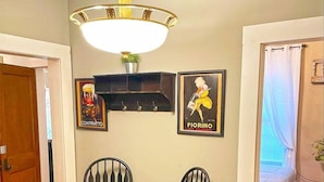 A room with two wooden chairs with checkered cushions, a wall-mounted shelf with two framed pictures, a hanging light fixture, and a door leading to another room. - King Charlie's Place in Historic Roscoe Village (Coshocton)