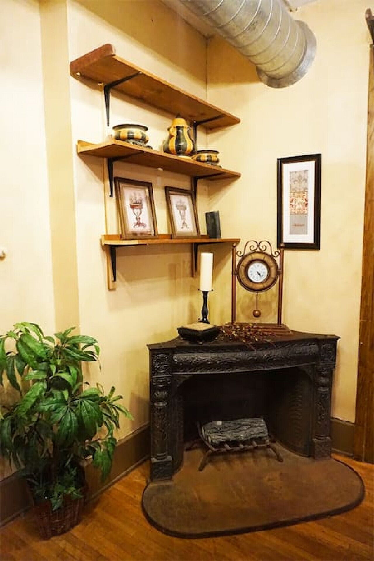 A corner room with a fireplace, wooden shelves with decorative items, a framed picture, and a potted plant.