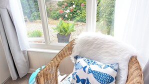 A wicker chair with a white furry throw and a seagull-patterned pillow, positioned by a window with a view of a garden.