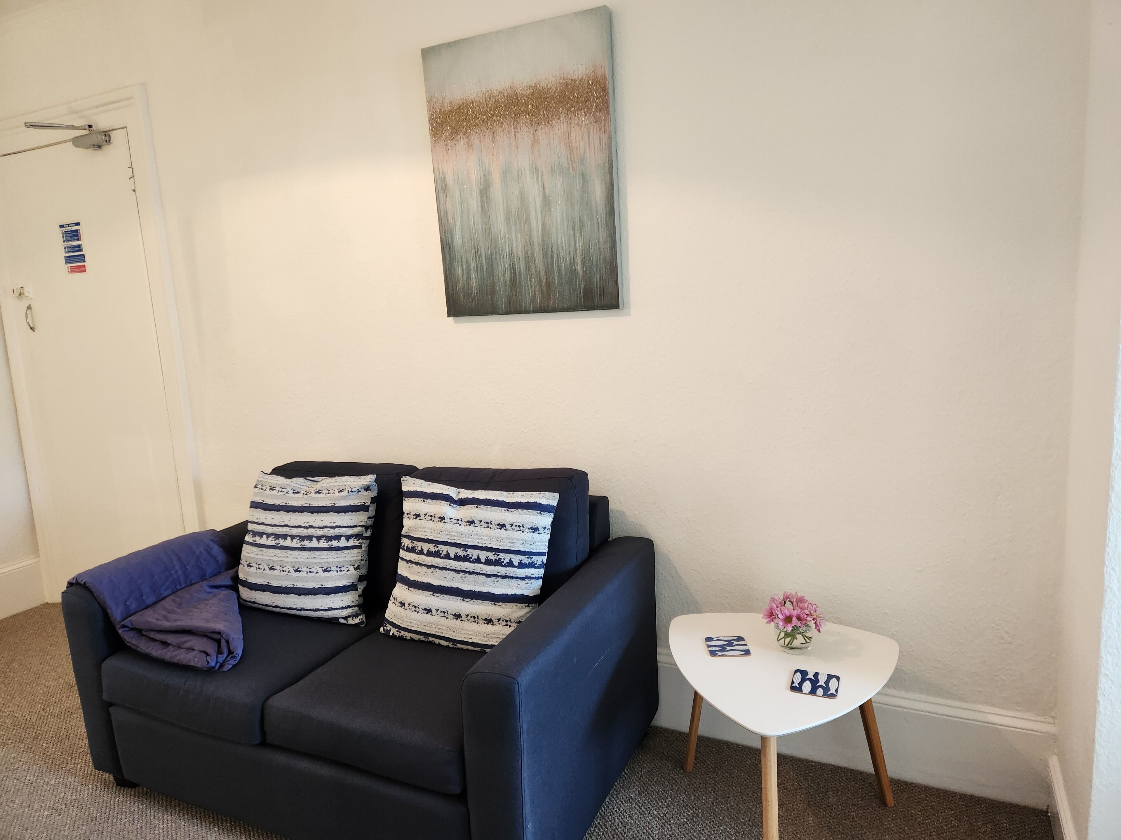 A living room with a dark blue sofa, two patterned cushions, a small white table with a vase of flowers, and a painting on the wall.