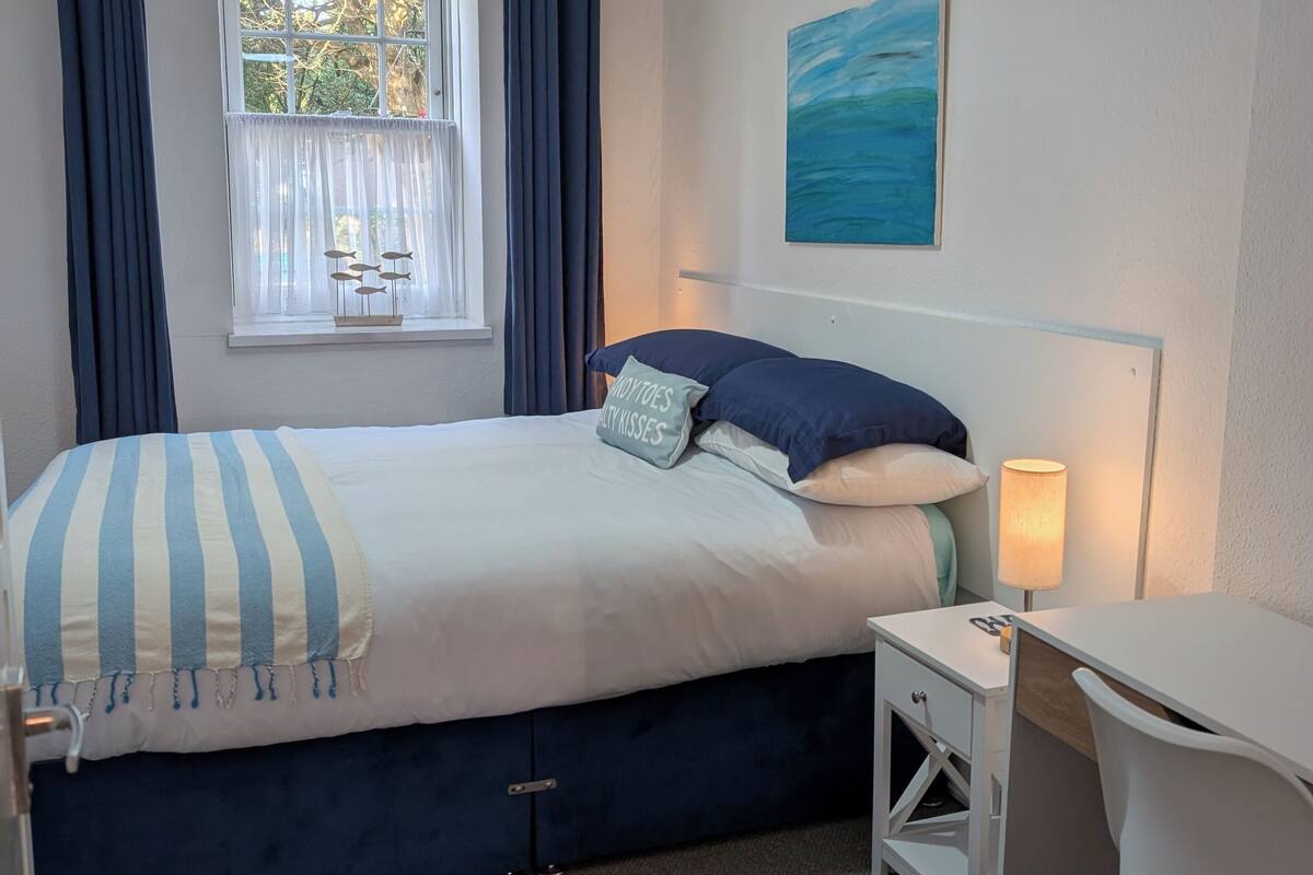 A bedroom with a bed, a desk, a chair, a window with curtains, a painting on the wall, and a hanging light fixture.