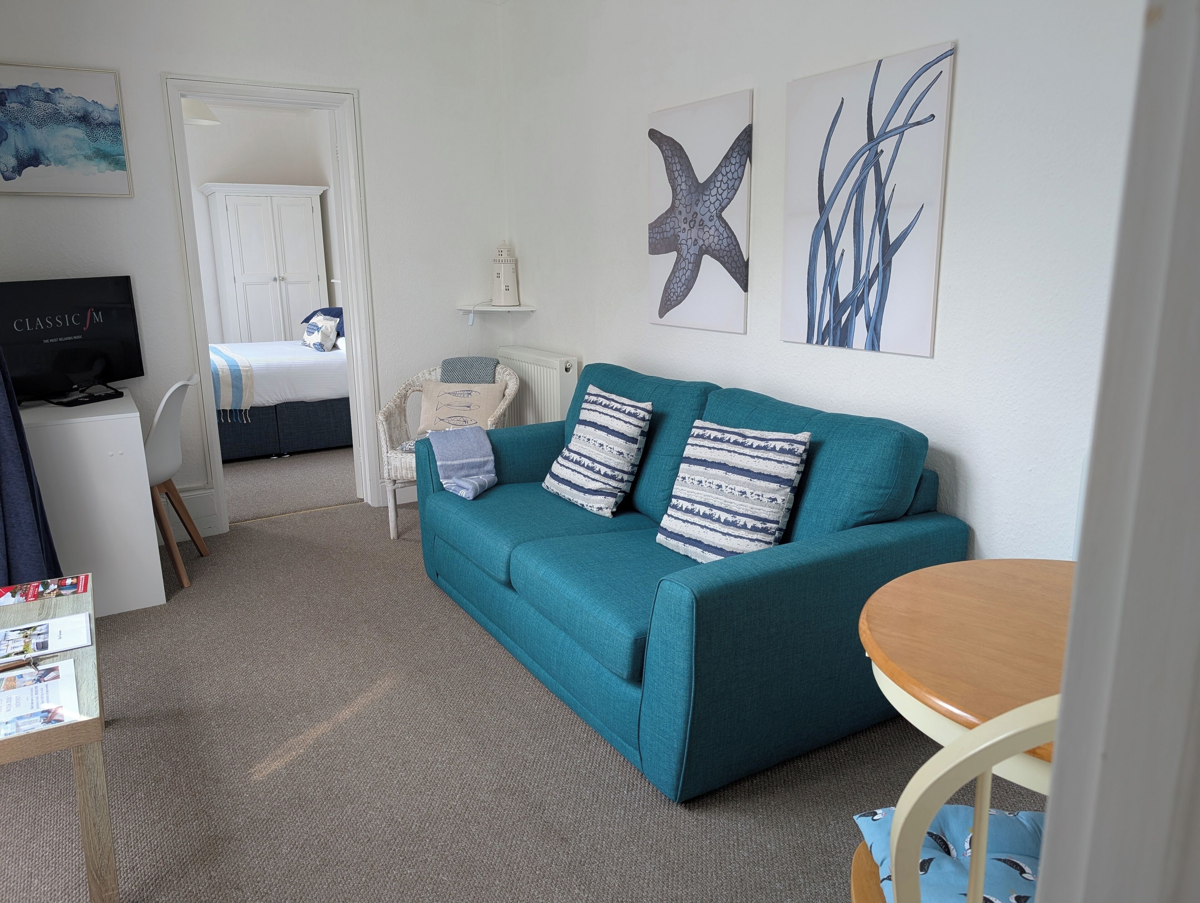 A living room with a teal sofa, a small round table, and a TV on a white cabinet. There are framed pictures on the wall, a wicker chair, and a bed in the background.