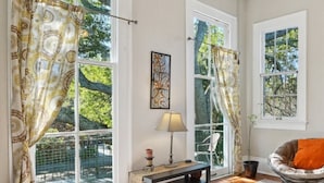 Une pièce dotée de grandes fenêtres, d’un ventilateur de plafond, d’une table avec une lampe et d’une chaise avec un coussin orange.