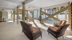 En udendørs overdækket terrasse med fletmøbler, en basketballkurv og en pool i baggrunden.