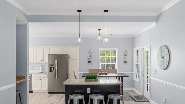 A modern kitchen with white cabinets, a stainless steel refrigerator, a central island with stools, pendant lighting, and a window with a view of greenery. - Pet-friendly Home w/ Pool, Hot Tub and Golf Cart (North Myrtle Beach)