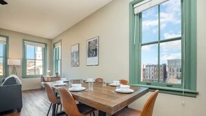 A dining area with a wooden table set for four, surrounded by chairs, with a city view through large windows. - The Penthouse- Heart of OTR w/Rooftop Terrace (Cincinnati)