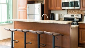 A kitchen with wooden cabinets, a stainless steel refrigerator, a microwave, a kettle, and a bar area with stools. - The Penthouse- Heart of OTR w/Rooftop Terrace (Cincinnati)
