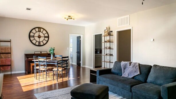 A spacious living room with a dark-colored sofa, a dining area with a wooden table and chairs, and a large wall clock.