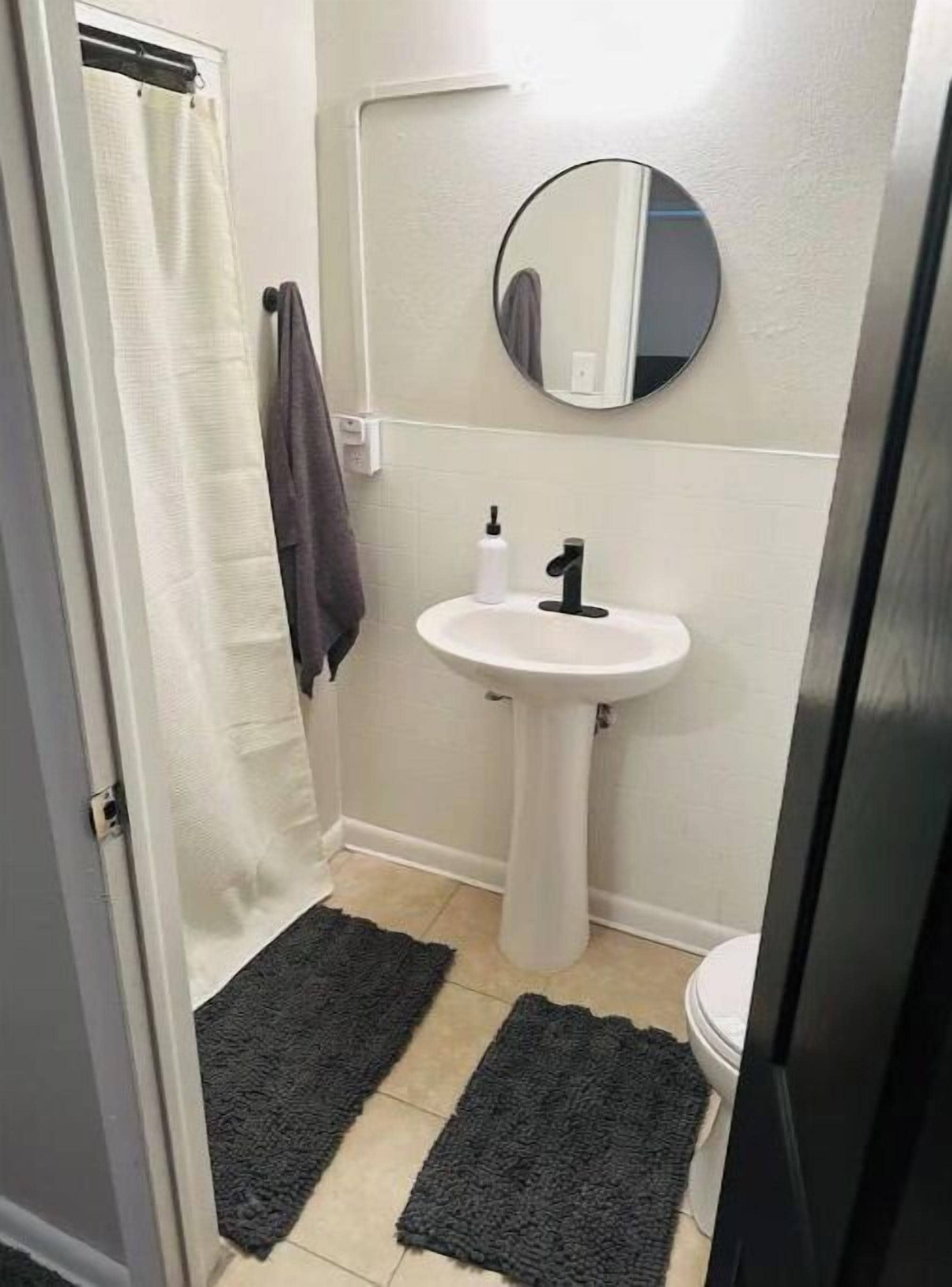 A small bathroom with a white sink, a mirror, and a toilet. There are two dark mats on the floor.