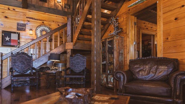 A rustic wooden interior with a leather sofa, ornate chairs, a wooden table, and a staircase.