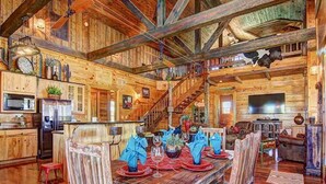 A rustic cabin interior with a wooden dining table set for four, a kitchen with modern appliances, and a staircase leading to an upper level.