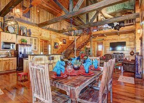 A rustic cabin interior with a wooden dining table set for four, a kitchen with modern appliances, and a staircase leading to an upper level.