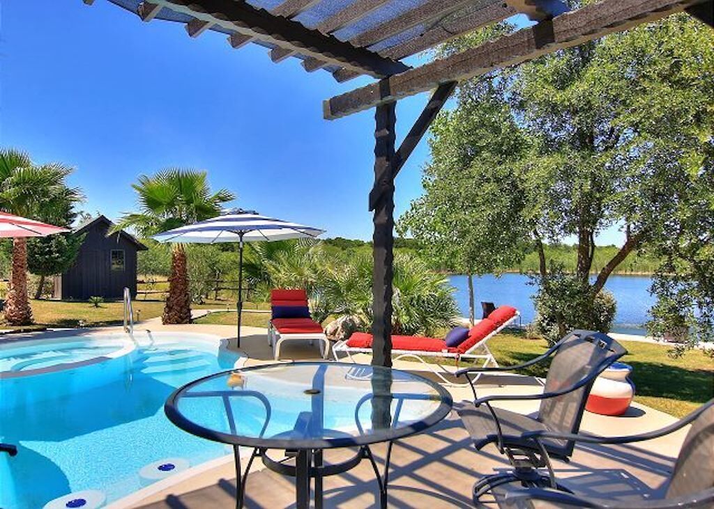 A pool area with lounge chairs and umbrellas, a pergola, and a view of a lake.