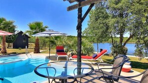 A pool area with lounge chairs and umbrellas, a pergola, and a view of a lake.