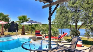 A pool area with lounge chairs and umbrellas, a pergola, and a view of a lake.
