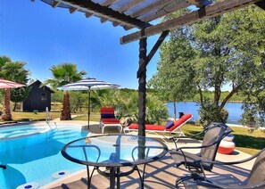 A pool area with lounge chairs and umbrellas, a pergola, and a view of a lake.
