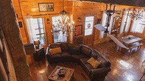 A cozy cabin interior with a wooden floor, a large L-shaped leather sofa, a chandelier, and a dining area with a table and chairs.