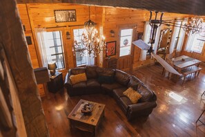 A cozy cabin interior with a wooden floor, a large L-shaped leather sofa, a chandelier, and a dining area with a table and chairs.