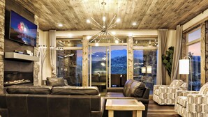 Una sala de estar moderna con techo de madera, televisor de pantalla plana, sofás de cuero, sillones estampados y vistas a montañas a través de puertas de vidrio.