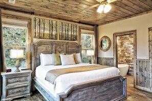 Quarto com cabeceira de madeira, uma cama grande com várias almofadas, mesa de cabeceira com abajur, espelho fixado na parede e parede com padrão de tijolos.