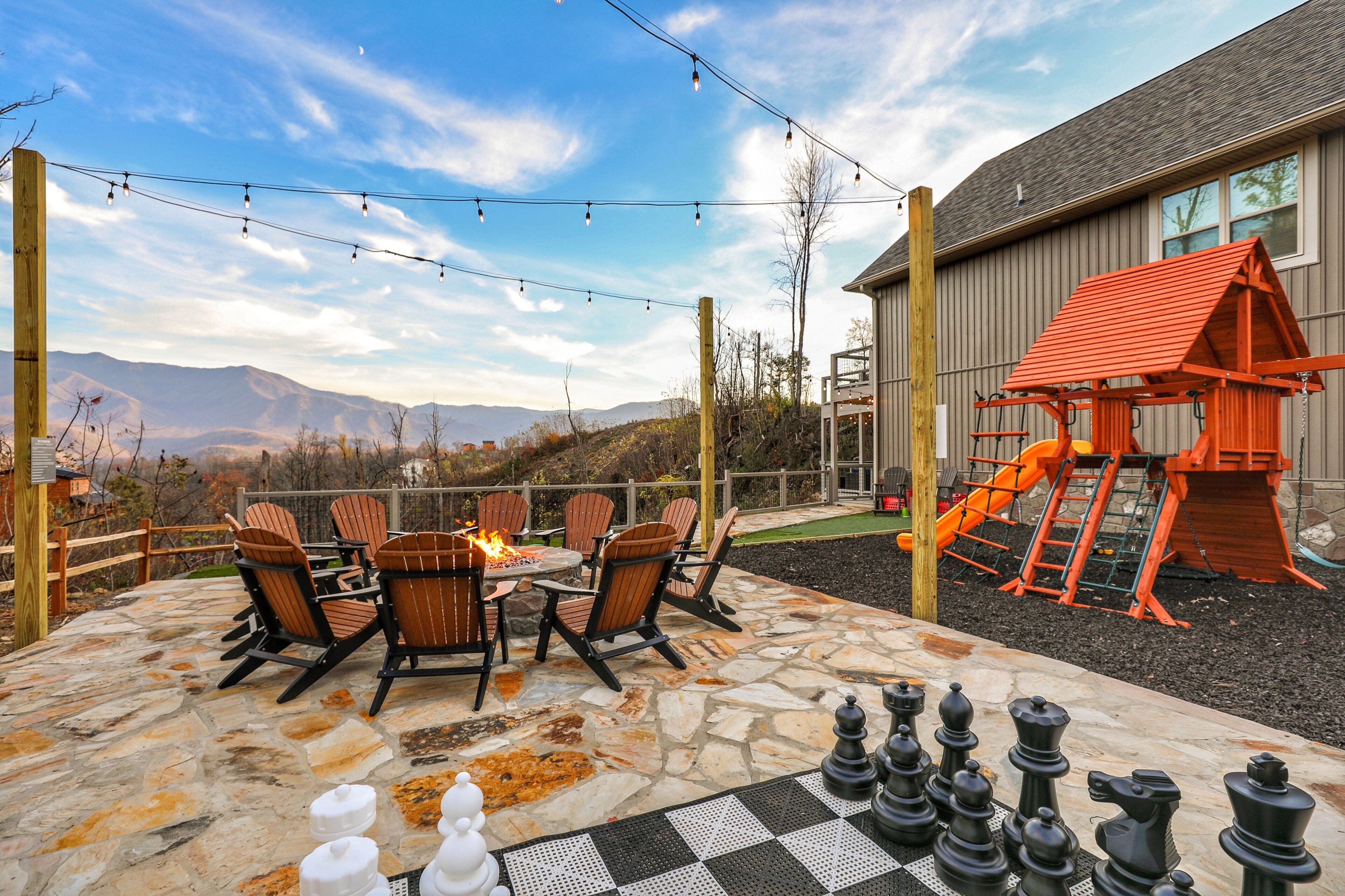 An outdoor patio with stone flooring, Adirondack chairs, a chessboard, and a playground structure.