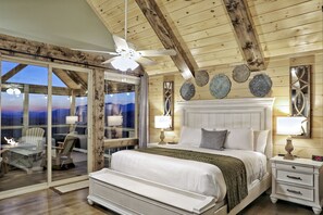 A bedroom with a large bed, two bedside tables with lamps, a ceiling fan, and a view through a glass door to the outside.