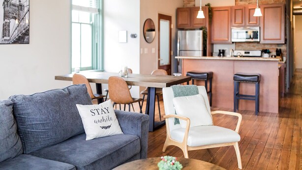 Un salon moderne avec un canapé gris, un fauteuil blanc et une table basse. Un coussin orné de l’inscription « RESTEZ un peu » est posé sur le canapé.