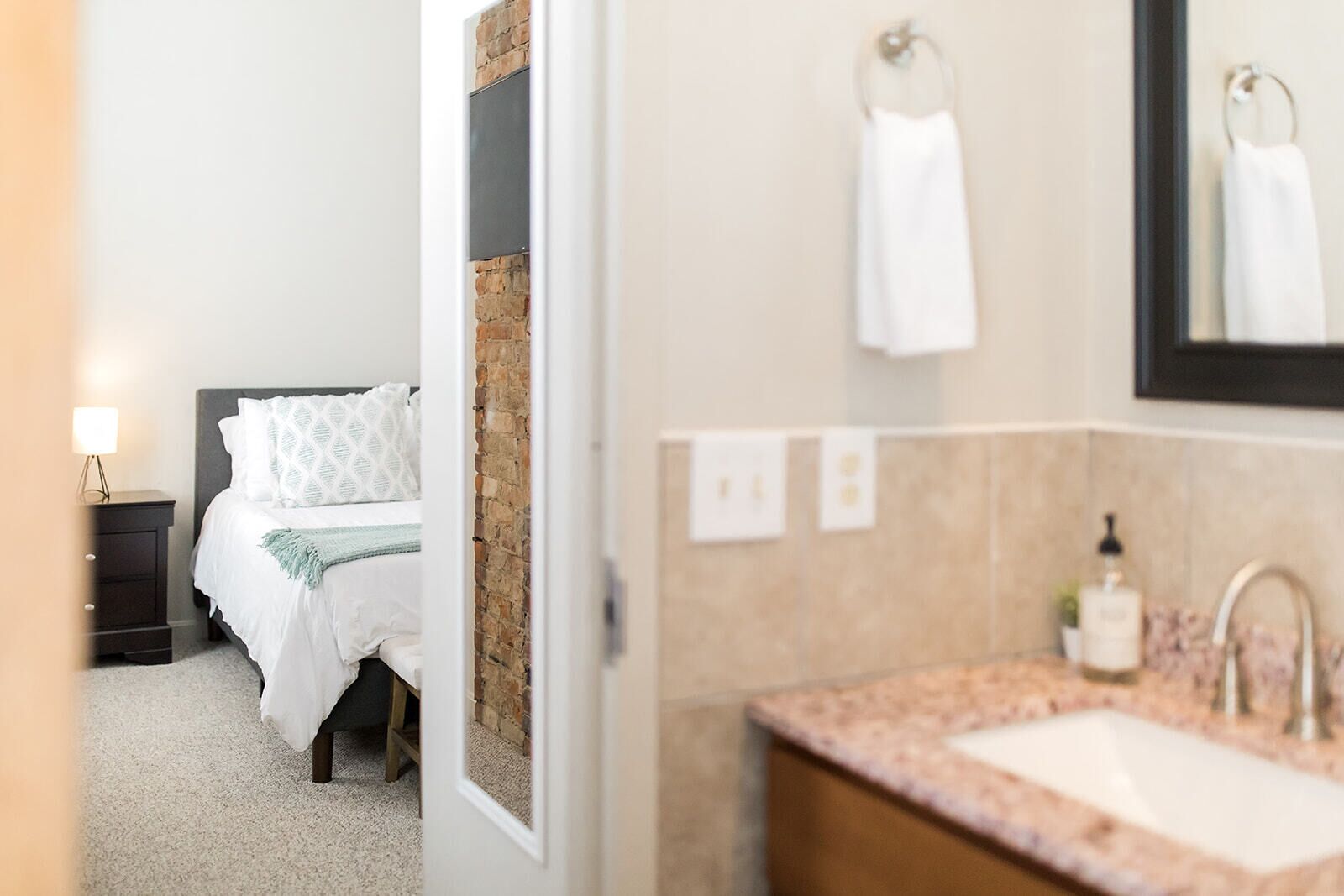 A hotel room with a bed, a nightstand, a lamp, a bathroom with a sink, mirror, and towel rack.