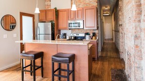 Une cuisine avec des meubles de rangement en bois, un réfrigérateur en acier inoxydable, un four à micro-ondes et une machine à café. On y trouve deux tabourets de bar et un luminaire suspendu.