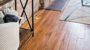 Une pièce avec un mur en briques, un plancher en bois, une petite table avec une plante, un panier blanc et une grande fenêtre.