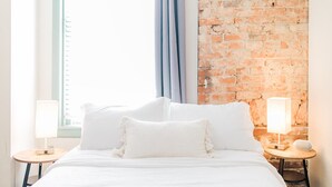 Un lit avec une literie blanche, deux tables de chevet avec des lampes, un mur en briques et une fenêtre avec des rideaux.