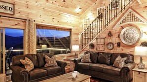A cabin interior with two leather sofas, a wooden coffee table, a staircase with a railing, and a large window with a view of the mountains. - EXTRAORDINARY Luxury View Cabin! Game Room! Theater! Porch w/ Fireplace! Hot tub (Gatlinburg)