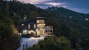 Una casa moderna de dos pisos con balcón, rodeada de un bosque denso.