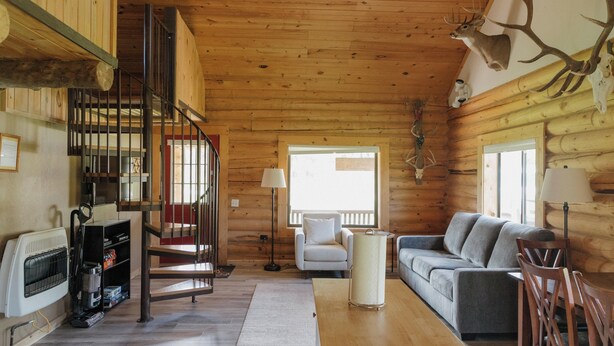 Interior de una cabaña de madera con una escalera, un sofá, una mesa y una cabeza de ciervo montada en la pared.
