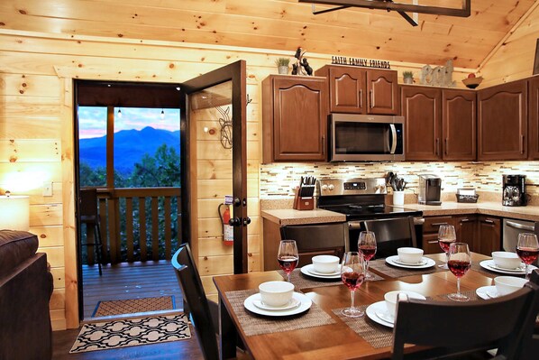 Interior de una cabaña de madera con comedor listo para una comida, una cocina con electrodomésticos modernos y vistas a montañas a través de la puerta abierta.