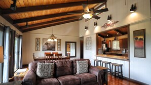 A spacious living room with a leather sofa, wooden ceiling beams, and a kitchen area visible through an open doorway.