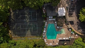 An aerial view of a property with a tennis court, a swimming pool, and a covered seating area.