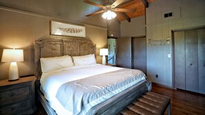 A bedroom with a large bed, wooden headboard, two bedside lamps, a ceiling fan, and a framed picture on the wall.