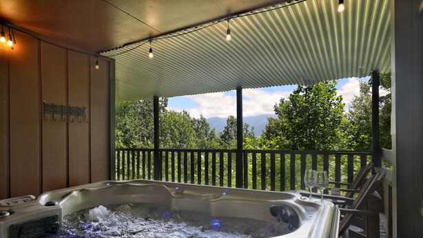 A hot tub with a view of trees and mountains, a wooden deck, and a metal roof.
